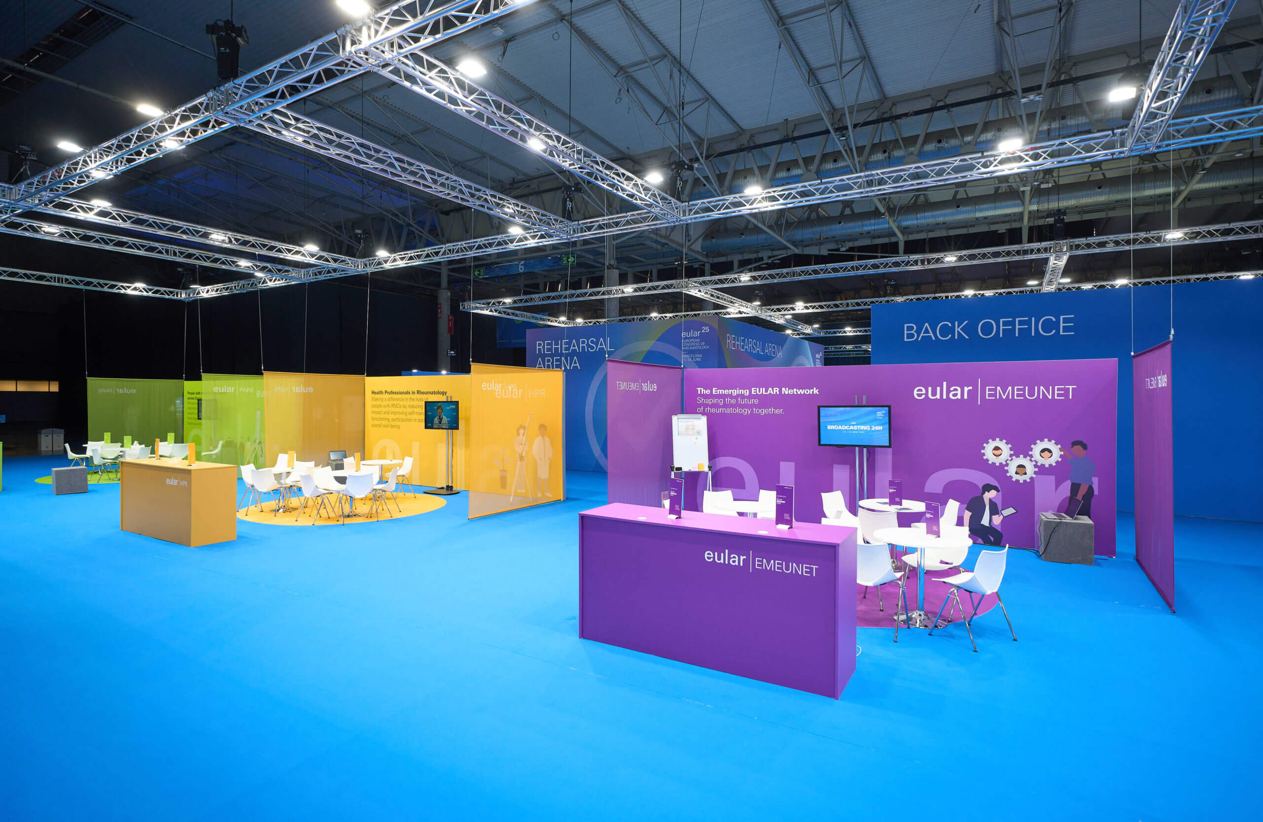 Área EULAR Village con mobiliario blanco, señalética de colores y pantallas para interacción en EULAR 2025.