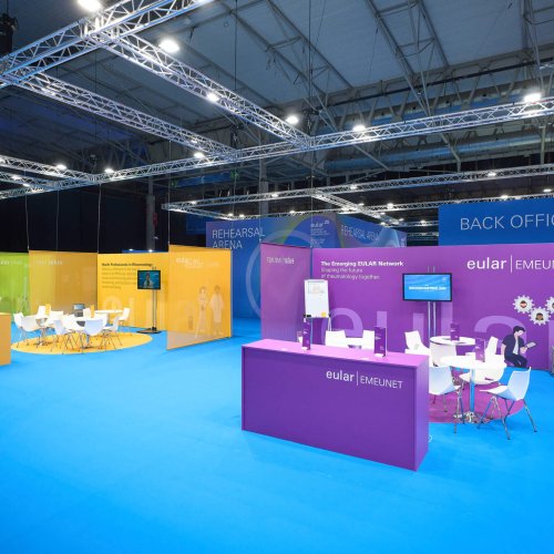 Área EULAR Village con mobiliario blanco, señalética de colores y pantallas para interacción en EULAR 2025.