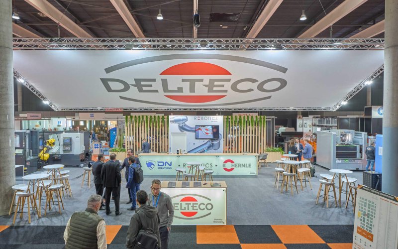 Vista frontal del stand de DELTECO en Advanced Factories 2025, con maquinaria en funcionamiento y zona de reuniones.
